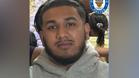 A man with black hair, a short black beard and thin moustache smiling and looking into the camera. There is a lilac flower wall behind him and his is wearing a thin, black headband to hold his hair out of his fa