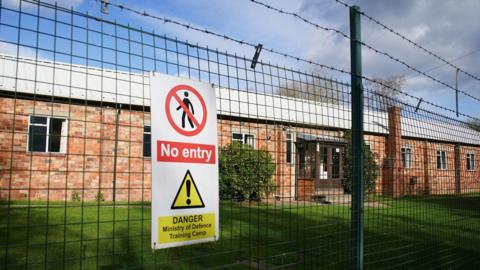 A general view of Crowborough Training Camp, East Sussex. There is a wire fence and a sign, with an area of grass behind it.
