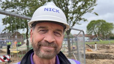 DIY SOS presenter Nick Knowles wearing a hard hat with the word: "Nick" on it and a purple hi-vis jacket. He is smiling with a brown beard and is stood in front of metal railings and work going on in some grass in the background. 