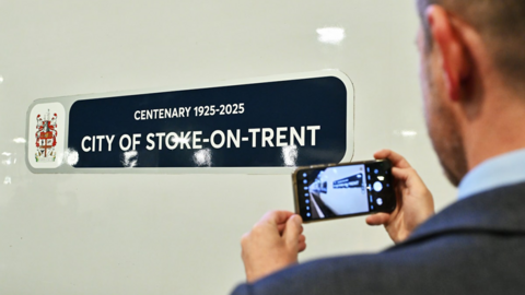 A close up of the nameplate on a train reading "Centenary 1925-2025 City of Stoke-on-Trent", with man taking a photo of the nameplate in the foreground on the right