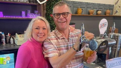 Claire in a bright pink top standing next to Neal in a pastel-coloured t-shirt, both behind a bar. Neal is pulling a pint.