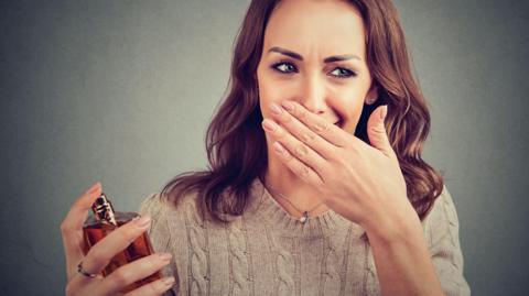 Woman holds a perfume sprayer in one hand with her other hand held to her face.
