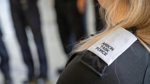 The shoulder of a woman who is looking away from the camera. She has blonde hair and the lapel on her jacket says, ARSON TASK FORCE.