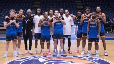 A group of 13 men all dress ing basketball blue and white clothing are stood in a row and smiling at the camera. Some are making peace sign gestures. They are stood on a basketball court.
