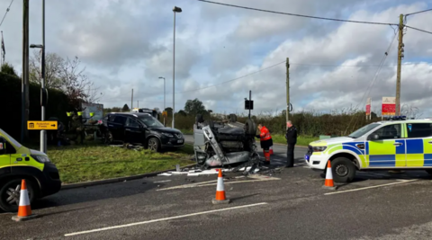 A police car and ambulance are parked nearby an upturned vehicle which is being attended by emergency service personnel.