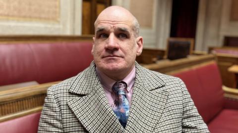 Leo looking at the camera. He is wearing a brown and cream checked jacket, a pink shirt and a blue patterned tie. He is sitting on a red bench.
