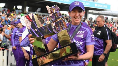 Northern Superchargers bowler Kate Cross smiles as she holds up The Hundred final after beating Southern Brave in the final