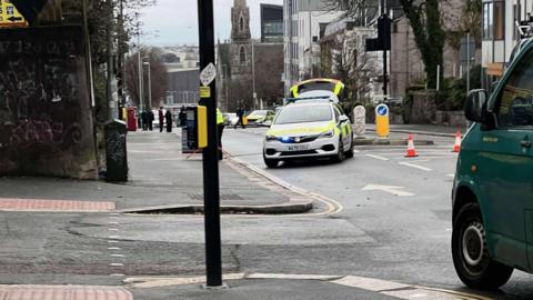 A police car is positioned at an angle blocking North Hill, Plymouth. A police officer is positioning road signs in the road. Orange cones block the other lane.