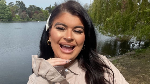 A woman is looking at the camera. She has one hand balanced on her chin and the other across her chest. She has a light cream top on and long dark hair. She is stood on the edge of a lake. She has an open mouth smile expression. 