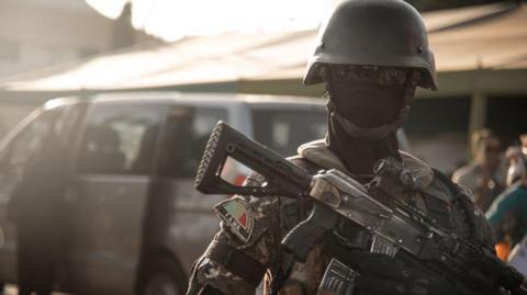 A member of the Malian military wearing a helmet, face mask and sunglasses looking into the camera and holding a rifle