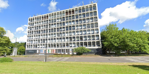 A high, brutalist building on the side of a main road. The sky is bright blue in the background and there are trees and grass around the building. There are several masts on top of the building. 