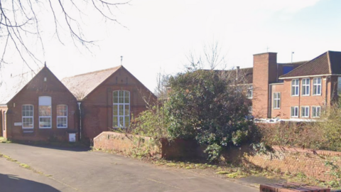 A Google street view image of the entrance to the car park of a former school. There are two building on the left of the image, a brick wall on which there is an overgrown bush in the middle of the image, and then a large building on the right of the image behind the bush.