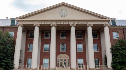 National Institutes of Health building pictured from the front. The building has several tall pillars and red brick walls.