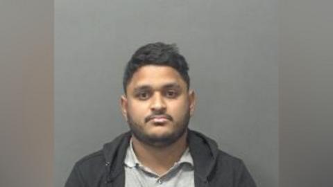 A mugshot of a man standing in front of a grey screen and wearing a hooded top.