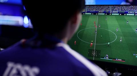 A person playing a football simulation video game on a large TV. The person has their back to the camera and the word Tess on the back of their shirt