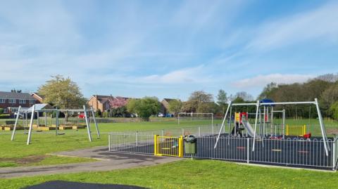 The park has a fenced off area which is part of wider playing fields. There are swings and a slide with a tower. Houses sit next to the edge of the lawns.