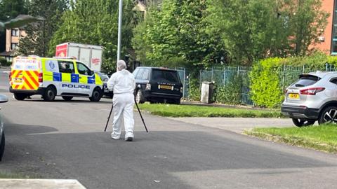 A forensics officer in white overalls on a street with a police van and parked cars 