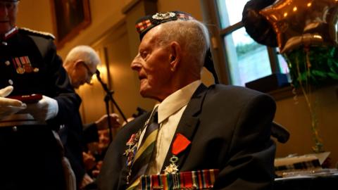 100-year-old veteran Jim Glennie sits wearing a suit, tie and military hat and medals.