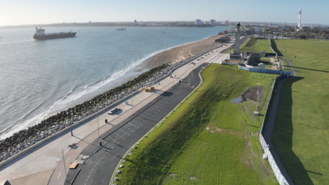 An aerial view of a stretch of coastline between the grassy Southsea Common, showing rock sea defences, a shingle beach, and the towering Portsmouth Naval Monument.