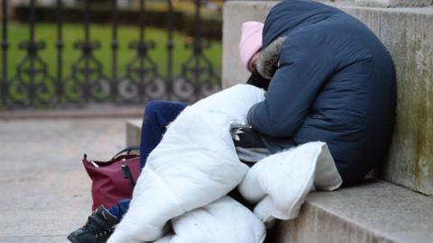 A person sat slumped over on a concrete step with their blue hooded coat covering their face as they sleep. They are holding a white duvet while sleeping next to someone else who wears a pink beanie hat, blue jeams and black nike trainers. There is a maroon handbag on the floor next to them