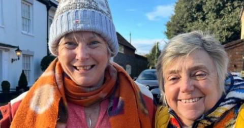 Two women standing by a road and smiling and looking straight at the camera. Caroline Giddings (left) is hearing a grey and blue hat, with a bobble on the top, a orange colourful scarf, and a pink top with a silver necklace round her neck. Denise Waters (right) has short grey hair, is hearing a colourful top with a yellow gilet. 