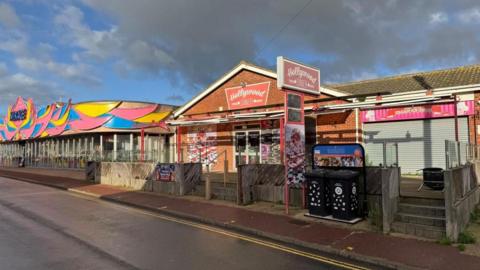 Hollywood Diner is a 1980s red brick building with a gabled end facing Beach Road, large windows, a canopy and signs outside, where there is a terrace outdoor dining area. The property is empty. Next to it is the brightly coloured facade of the Mirage amusement arcade, which has a wraparound glass door.