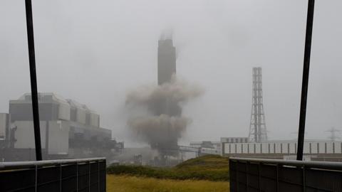 Smoke surrounds the a tall chimney as it is demolished. It is a grey, foggy day with a white sky behind the chimney. A building can be seen to its left-hand side. 