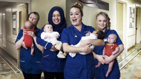 Four female midwives are standing together and smiling as they each hold a baby. They are in a hospital corridor, with a sign behind them saying 'delivery suites'.