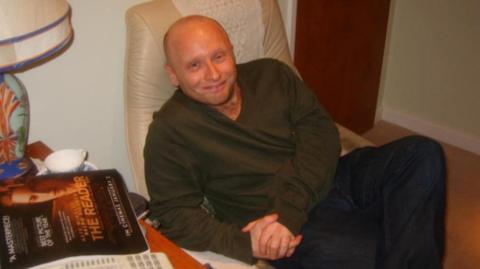 Dan Kay smiles at the camera while sitting in a leather armchair. He is bald and is wearing a green sweatshirt and jeans