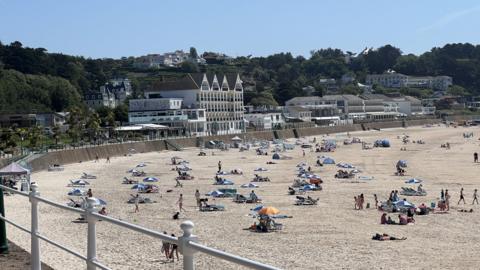 A view of a light. sandy beach on Jersey. There's people sat on the beach on blankets and umbrellas. There are buildings lining the seafront as well as trees surrounding them. 