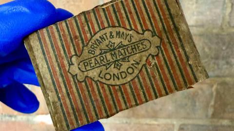 A box of matches, with red, black and cream stripes, with writing in the middle. A person is holding them up, wearing blue gloves. 