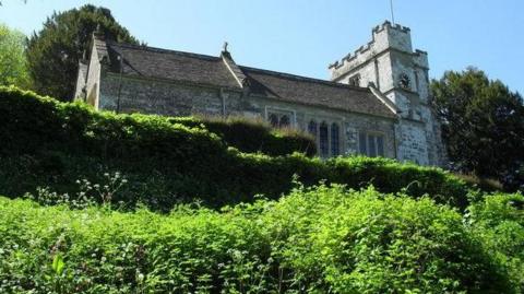 Looking up the escarpment towards the medieval stone church with clock tower with bushes and a hedge in the foreground