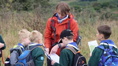 A scouts leader giving instructions to four younger members at a field. They are wearing backpacks and blue and white scarves. 