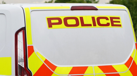 The image shows the rear of a British police van. The van has high-visibility markings in red and yellow chevrons. The word “POLICE” is printed in large, bold red letters on a yellow background near the top of the rear door.