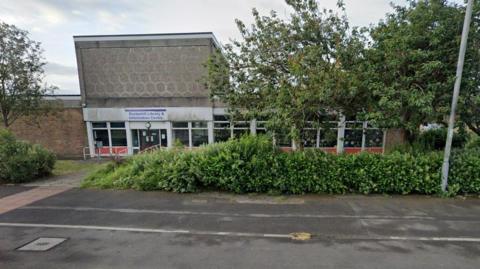 Eccleshill Library, a brutalist building with a sign indicating its name above the front entrance. Trees and shrubs are in the foreground, in front of the building itself.