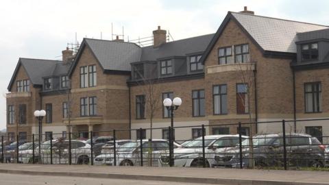 A modern brick care home with large windows and dark slate-style roofing. Several cars are parked in front of the building and are visible behind a black metal fence.