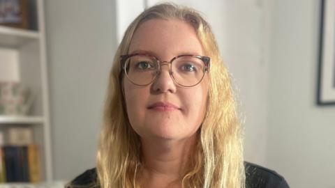 Emily Barley is sitting on a sofa in her lounge and she is holding a piece of paper which has two small handprints printed on it. Ms Barley has long blonde hair and is wearing glasses and a black top. There are framed photographs on the shelves to her right.