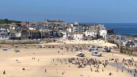 A beach in Cornwall. There are hundreds of people on the beach and the sky is blue. There are boats dotted across the beach. Houses sit behind the beach and buildings.