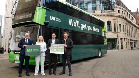Four people stand in front of a green double-decker bus with buildings behind. The people are holding two banners that read 'Weaver Network' and 'Better buses for West Yorkshire'. The sign on the side of the bus reads 'Weaver Network: We'll get you there'.