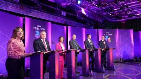 Two women and four men stood in front of a podium each, in front of purple BBC election branding