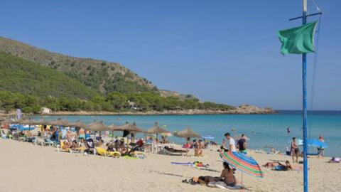 A sandy beach with sunbathers, swimmers, a green flag on a pole and straw beach umbrellas. A rocky spur is in the background at the far end of a bay.