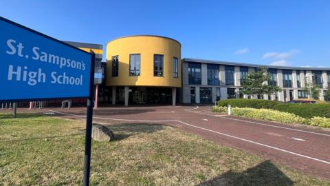 A modern-looking school with colourful, yellow walls and curves. A sign says St Sampson's School.