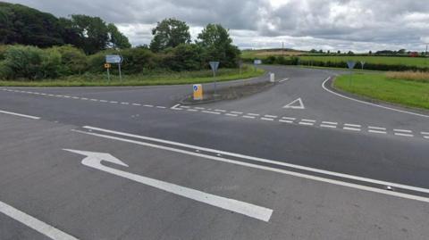 Streetview of a junction, with a large white arrow in a turning lane in the middle of the road pointing towards a road heading off round a corner. There is a yellow and white traffic cone at the mouth of the junction and a sign pointing up the road. Green trees and fields are beyond.