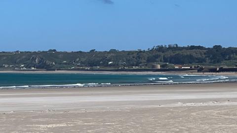 A picture of St Ouen in Jersey. It is a sandy beach and in the distance there is blue water. In the distance there is green land with buildings along the shore.