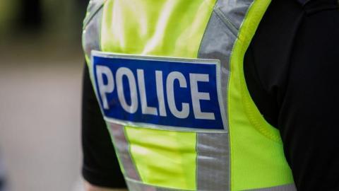 The back of a police officer wearing a yellow police jacket with a blue badge that says police in white letters.