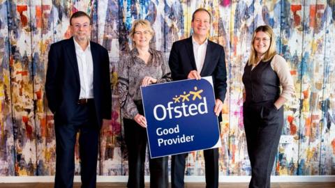 Two men and two women stand in front of a sign that says "Ofsted good provider" - the men are wearing suits with no ties. The women are wearing smart trousers.