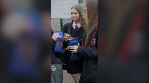 Two female pupils stand holding blue pouches containing their mobile phones.