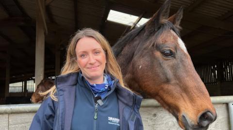 Emma Carter is standing next to a brown horse. Emma is wearing a blue navy jacket and has long brown hair. To the right is a brown horse with a white spot on its head. Its ears are pointing forwards.