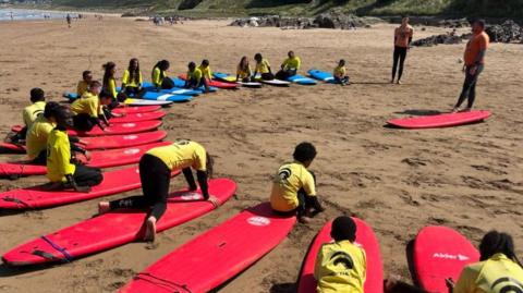 A group of 19 children sit on red and blue surfboards on a beach while having surfing lessons in Devon. Two men are instructing them.