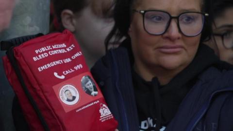 A woman wearing black-rimmed glasses holds a red package marked "bleed control" kit and which also bears a picture of Harvey Willgoose